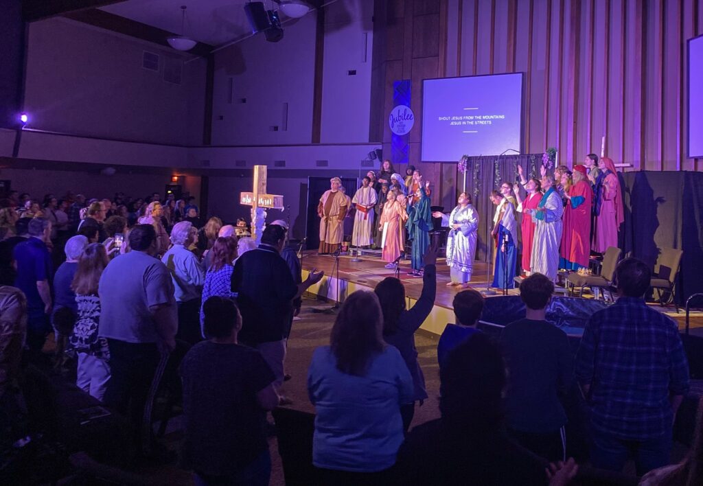 A group of people, dressed in costumes, perform on a stage in front of an audience inside a church. A wooden cross stands prominently at the forefront, partially obscuring stage view.