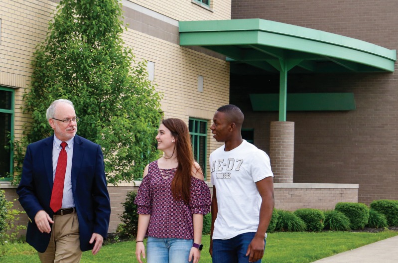 Three people are walking and conversing outside a building with green trim. A man in a suit and tie is accompanied by two young adults, one in a patterned blouse and jeans, and the other in a white T-shirt.