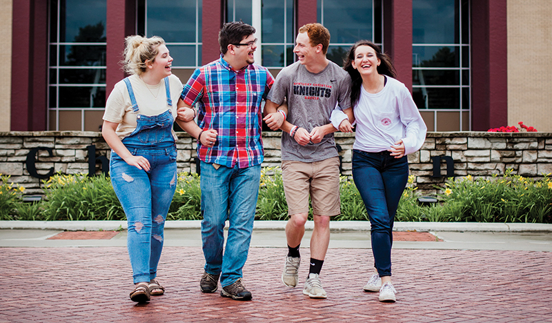 Four people walking and laughing together outside, with a brick building in the background. Two on the left wear casual outfits, and two on the right wear t-shirts and jeans.