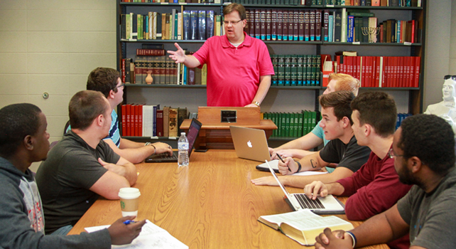 A man in a red shirt is speaking at a lectern in front of a table with seven seated individuals. Bookshelves filled with books are in the background.