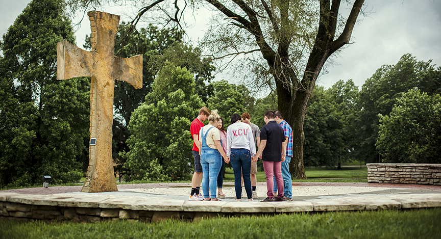 A group of five people stands in a circle holding hands near a large stone cross outdoors, surrounded by trees and greenery.