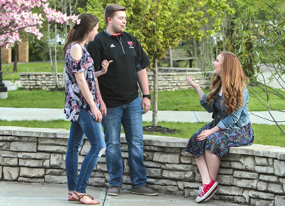 Three people, two standing and one sitting on a stone ledge, engaging in conversation outdoors near blooming trees and greenery.