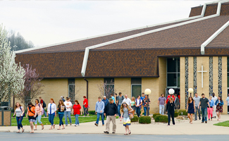 A group of people, including men, women, and children, exit a modern church building with brown roof and cross symbol in an outdoor setting. Trees and shrubs decorate the surroundings.