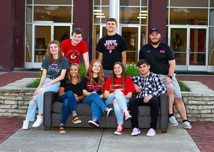 Eight young adults are sitting on and around a couch outdoors, all smiling. They are dressed casually, and a building with large glass windows is visible in the background.