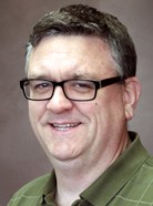 A man with short hair wearing glasses and a green collared shirt, smiling at the camera against a plain background.