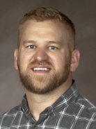 A man with short blond hair and a beard is wearing a gray checked shirt and smiling at the camera against a gray background.