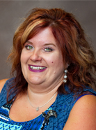 A woman with shoulder-length wavy hair dyed red and blonde, wearing a blue patterned top, smiling, and looking towards the camera with a plain background.