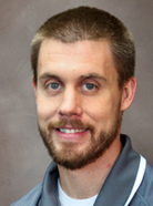 A man with short brown hair, a beard, and a mustache is wearing a gray collared shirt and smiling at the camera against a neutral brown background.