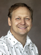 A man with short hair and wearing a white shirt with small green and red prints smiles at the camera. He has a light complexion and the background is brown.