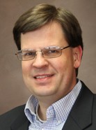 A man with glasses, short brown hair, and wearing a suit jacket and checkered shirt, smiling against a plain brown background.