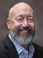 A bald man with a gray beard, wearing a checkered shirt and dark blazer, smiles against a plain, dark background.