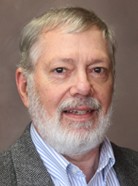 A man with gray hair and a beard is wearing a gray suit jacket over a blue and white striped shirt. He is posing in front of a neutral background.