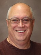 A bald man with glasses is smiling at the camera. He is wearing a dark-colored sweater. The background is plain and neutral.