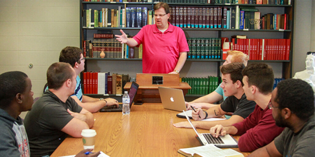 A person in a pink shirt stands at a lectern, speaking to a group of six seated people. The group is gathered around a table with books, laptops, and notebooks, in a room with shelves of books.