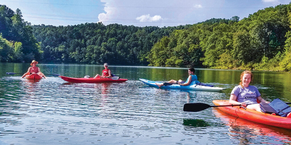 Four people paddle kayaks on a calm lake surrounded by lush, green trees on a sunny day.