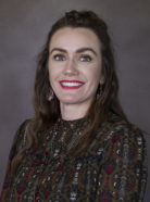 A woman with long brown hair wearing a patterned blouse smiles at the camera against a plain background.