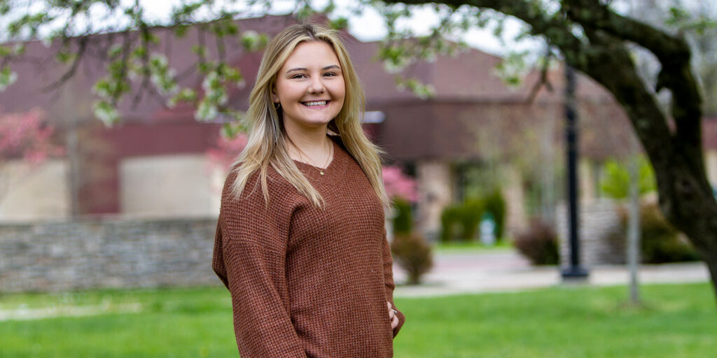 A young woman in a brown sweater stands outdoors on a grassy area, smiling. Blurred buildings and trees with small flowers are visible in the background.