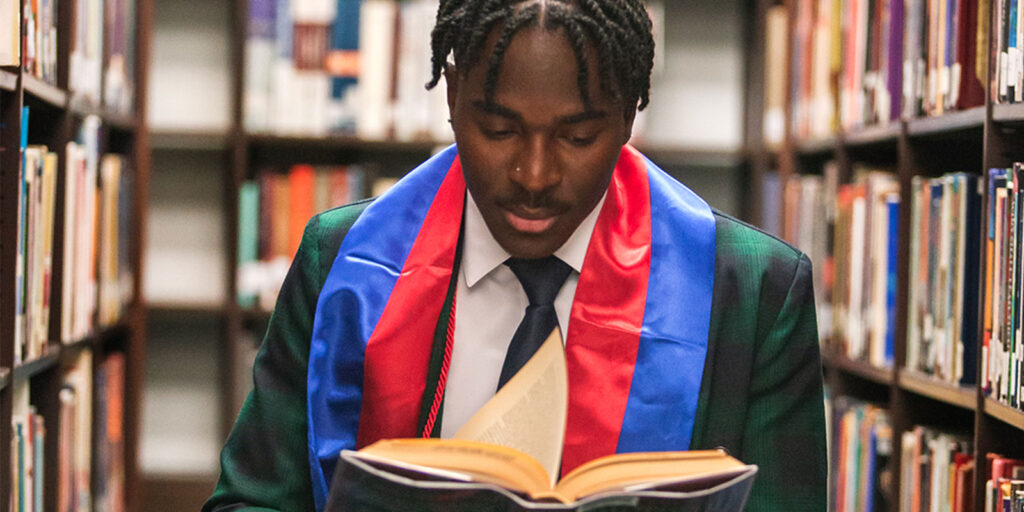 A person wearing a graduation stole reads a book in a library aisle.