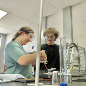 Two individuals wearing safety goggles conducting an experiment together on a lab bench in a science classroom.