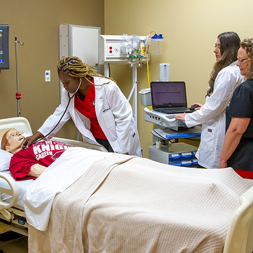 Three healthcare professionals practice medical procedures on a patient simulator in a hospital room. One uses a stethoscope, another operates a computer, and the third oversees the activities.