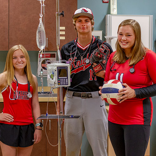 Three young adults in sports attire stand in a medical setting. One holds a volleyball, another a baseball glove, and the last one has an IV drip setup beside them.