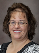 A woman with shoulder-length brown hair and glasses is smiling at the camera, wearing a black and white patterned blouse against a plain background.