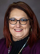 A woman with shoulder-length brown hair, glasses, and hoop earrings. She is smiling and wearing a dark blouse with a purple jacket.