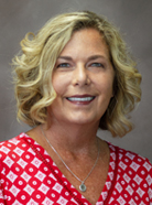 A woman with shoulder-length blonde curly hair, wearing a red and white patterned top, smiles in front of a gray background.