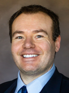 A man with short brown hair smiling, wearing a dark suit, light blue shirt, and blue tie.