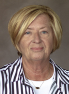 A woman with short blonde hair wearing a white top layered with a black and white striped shirt. She is looking directly at the camera against a neutral background.