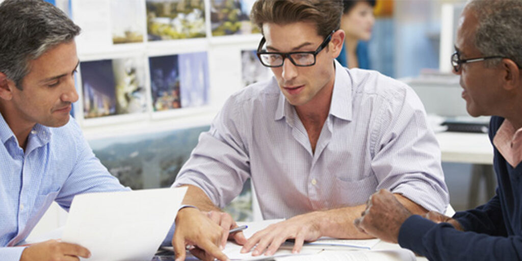 Three men are sitting at a table, engaged in a discussion. They are looking at documents and one of them is pointing at a sheet of paper. The background shows blurred office desks and photos on the wall.