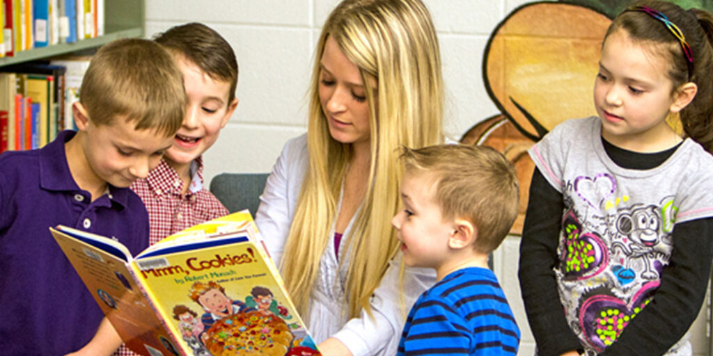 A young woman is reading a book titled "Warm Cookies!" to four young children, who are gathered closely around her, in a library setting.