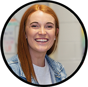 A person with long red hair, smiling, wearing a denim jacket over a light-colored shirt.