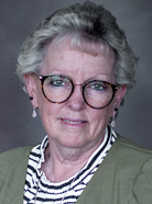 Portrait of an older person with short gray hair, glasses, and a striped shirt underneath a green cardigan, against a gray background.