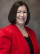 A woman with shoulder-length dark hair is smiling and wearing a red blazer over a black top. She stands against a neutral background.