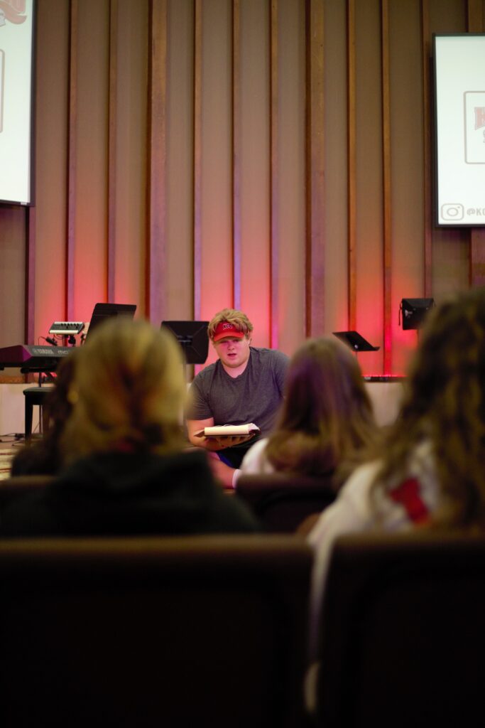 A person wearing a red visor speaks to an audience in a dimly lit room with red backlighting and vertical wooden panels.