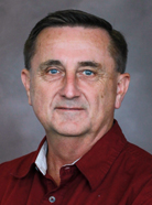 A man with short hair wearing a maroon shirt and white undershirt looks directly at the camera against a plain background.