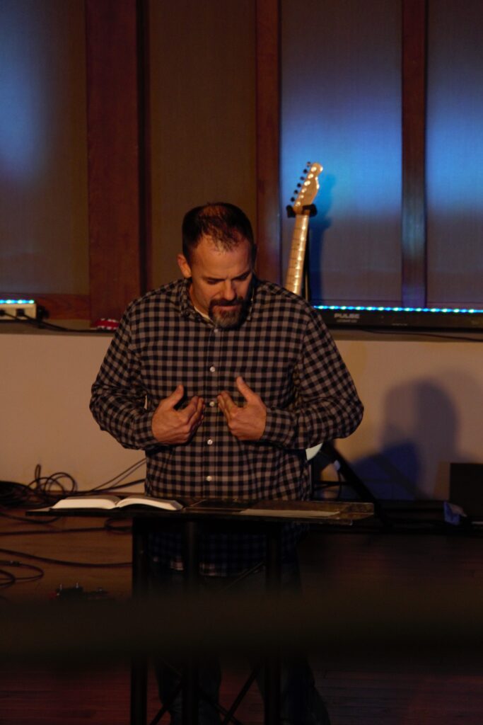 A man stands behind a podium, gesturing with his hands. An open book is on the podium, and a guitar is in the background.