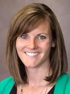 A woman with brown hair and blue eyes, wearing a dark top and a green jacket, smiles at the camera against a neutral background.