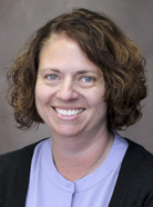 A woman with curly brown hair, wearing a black sweater and a light purple shirt, smiles at the camera against a plain background.