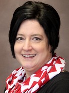 A person with short dark hair, wearing a red and white patterned scarf and black clothing, smiles at the camera in front of a plain background.