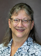 A woman with glasses, gray hair, and wearing a patterned shirt smiles in a headshot against a plain background.