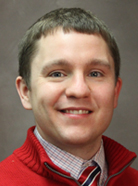 A man with short brown hair is smiling, wearing a red sweater over a patterned shirt and striped tie. He is posed against a plain gray background.