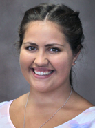 A smiling person with dark hair pulled back, wearing a light-colored top and a necklace, against a neutral background.