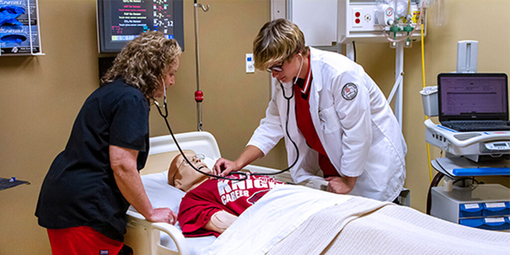 Two healthcare professionals examine a medical mannequin in a clinical setting, using stethoscopes and medical equipment to simulate patient care.