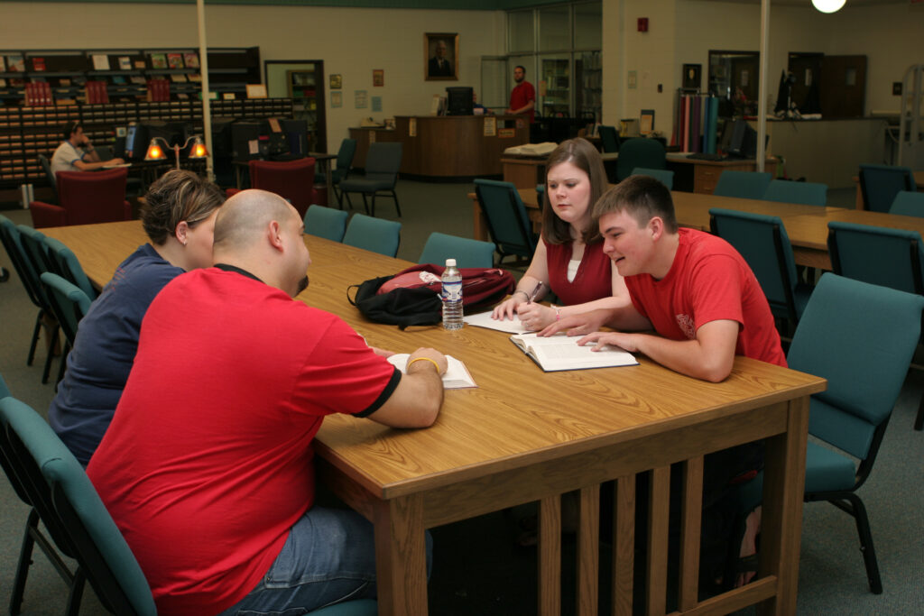 Four people are seated around a wooden table in a library, engaged in discussion. One man, holding a pen, points to a notebook. Others listen attentively. Shelves and desks are visible in the background.