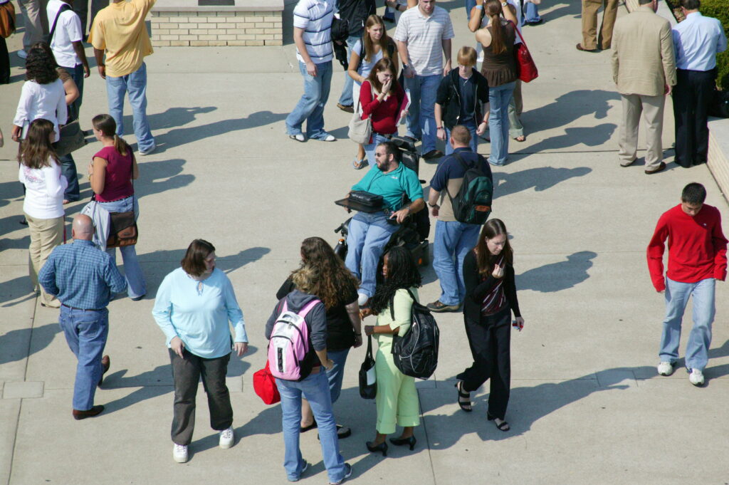 A diverse group of people, including students with backpacks and a person in a wheelchair, are mingling and walking in an outdoor paved area surrounded by buildings.