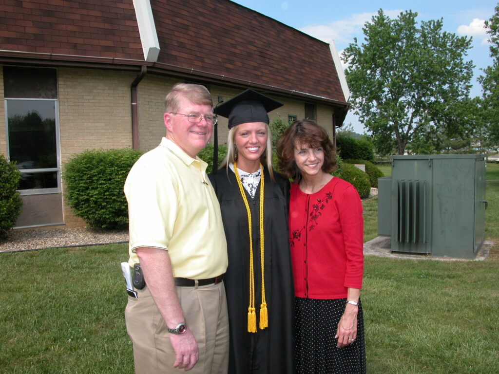 A graduate in cap and gown stands between an older man in a yellow shirt and a woman in a red cardigan in front of a building and grassy area.