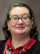 A woman with glasses and shoulder-length hair smiles. She wears a red and black outfit with a patterned scarf and pearl earrings against a plain background.