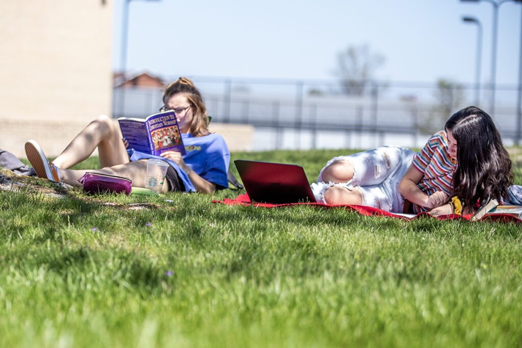 Two people lying on the grass; one is reading a book and the other is using a laptop.